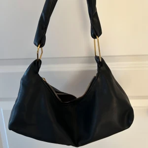 Svart skinnväska med gulddetaljer - Snygg svart axelväska i skinn med guldfärgade detaljer. Väskan har en  dragkedja och en bekväm axelrem:)