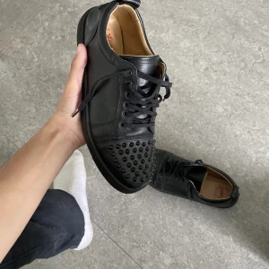 Loubution skor  - Säljer ett par svarta sneakers i skinn med coola nitar på tån. De har en röd sula som ger en snygg kontrast och snörning för perfekt passform. Perfekta för att ge din outfit en edgy touch! 