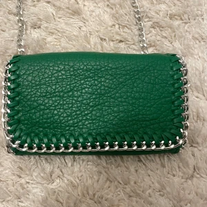 Grön axelväska med kedja från Tiamo - Snygg grön axelväska med silverkedja Perfekt för en kväll ute eller en dag på stan. Väskan har en praktisk dragkedja och magnetknappar för säker förvaring. Den är både stilren och funktionell!