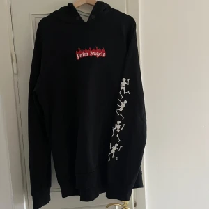 Palm Angels Hoodie - Säljer en svart hoodie från Palm Angels med coola skelettmotiv längs ärmen och ett rött logotryck på bröstet. Den är gjord av 100% bomull och är i storlek L, modellen är oversized så passar även större storlekar. Hör av er vid frågor eller kom med ett prisförslag!