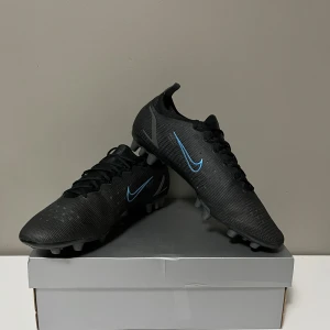 Nike Mercurial Vapor 14 AG - Snygga svarta fotbollsskor från Nike med blå detaljer och snörning. Skorna har en slitstark sula med dobbar för optimalt grepp på planen. Perfekta för fotbollsspelare som vill ha stil och funktionalitet. 9/10 skick använda endast 1 gång (priset kan diskuteras!)