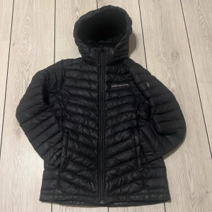 Svart dunjacka från Peak Performance - Säljer en svart dunjacka från Peak Performance. Jackan har en praktisk huva och dragkedja framtill. Perfekt för kyliga dagar med sin quiltade design som håller värmen. Skick 8/10