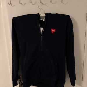 Marinblå kofta från Comme des Garçons (Play) - Säljer en stilren marinblå kofta från Comme des Garçons( Play) med det ikoniska röda hjärtat på bröstet. Tröjan har dragkedja och huva, perfekt för en casual look. Passar utmärkt till både jeans och joggers.