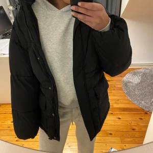 Svart pufferjacka med luva  - Snygg och varm svart pufferjacka perfekt till vintern med en snygg och stor luva. Fickor med knappar framtill, och en dragkedja som går ända upp till halsen. Resår i midjan som man kan dra åt om man vill få jackan tightare i midjan. Helt ny, aldrig använd och med prislappen på! I storlek 34.