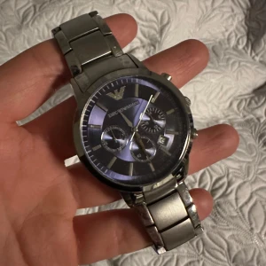 Armani klocka - Säljer en stilren och elegant klocka från Emporio Armani i silverfärg. Den har en rund urtavla med flera små urtavlor och ett datumfönster. Armbandet är i metall. Säljer billigt eftersom den har slutat ticka.