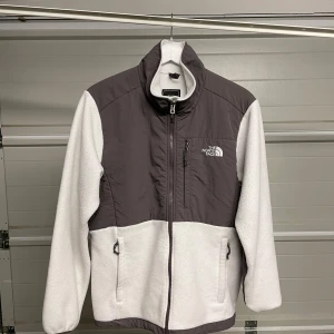 Vit och grå fleecejacka från The North Face - Säljer en supermysig fleecejacka från The North Face i vitt och grått. Den har en dragkedja framtill och två praktiska fickor med dragkedja. Perfekt för kyliga höst- och vinterdagar. Jackan har en hög krage för extra värme och komfort. Säljes i befintligt skick. Storlek: L