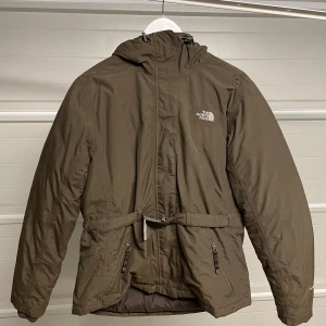 Brun jacka från The North Face - Säljer en brun jacka från The North Face i mycket bra skick. Den har en praktisk huva och dragkedja framtill. Jackan är perfekt för höst och vinter, med en normal passform och långa ärmar. Den har också ett bälte i midjan för extra stil och funktion. Säljes i befintligt skick. Storlek: L