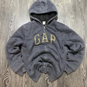 Mörkgrå zip hoodie från GAP - Säljer en mörkgrå hoodie från GAP med dragkedja och stor logga på bröstet. Hoodien har en mysig fleecefodring och ribbade muddar vid ärmslut och nederkant. Perfekt för kyliga dagar! Priset kan diskuteras!