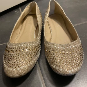 Glittriga ballerinaskor med pärlor - Snygga ballerinaskor i guld med glittrande detaljer och pärlor. Perfekta för att ge en extra touch till din outfit. Skorna har en rund tå och är dekorerade med små paljetter och pärlor för en elegant look.