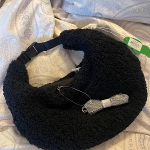 Svart axelväska från Nunoo - Snygg och trendig svart axelväska från Nunoo i ett fluffigt material. Väskan har en justerbar rem och en praktisk dragkedja för säker förvaring. Perfekt för att ge din outfit en mysig touch. Nypris 899:-