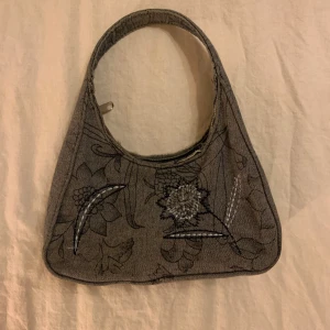 Grå axelväska med blommigt mönster - Säljer en unik grå axelväska med ett vackert blommigt mönster. Väskan har en dragkedja upptill och är perfekt för både vardag och fest. Den är i bra skick och har en stilren design som passar till många olika outfits.