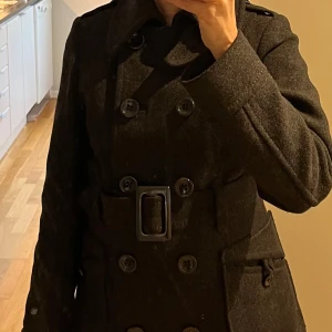 Mörkgrå kappa i ull från H&M - Säljer en mörkgrå kappa från H&M i storlek 34. Den har en klassisk knäppning och ett snyggt bälte i midjan för en figursydd look. Perfekt för vinter med sina långa ärmar och stilrena design. Material 80% och polyamid 20%. Fodret är polyester. 