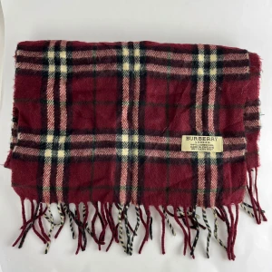 Rutig halsduk från Burberry - Säljer en klassisk rutig halsduk från Burberry i vinrött, svart och vitt. Den är gjord av mjuk ull och har fransar i ändarna. Perfekt för att hålla sig varm och stilig under kyliga dagar. Passar både höst och vinter!