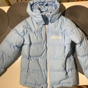 Ljusblå dunjacka från Hoodrich - Säljer en snygg ljusblå dunjacka från Hoodrich med dragkedja och huva. Jackan har en broderad logga på bröstet och ryggen, vilket ger en cool touch. Perfekt för kyliga dagar.