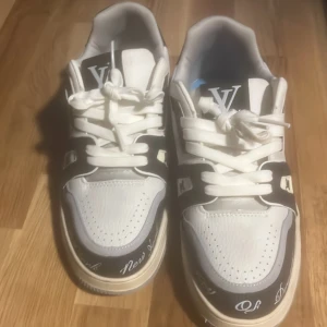 Vita och svarta sneakers från Louis Vuitton - Säljer ett par snygga Louis Vuitton sneakers i vitt och svart. De har en cool design med LV-loggan och textdetaljer på sidan. Skorna har snörning och en bekväm sula med mönster. Perfekta för att ge din outfit en lyxig touch!