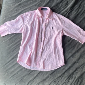 Rosa skjorta från Ralph Lauren - Säljer en snygg rosa skjorta från Ralph Lauren i storlek XL. Skjortan är långärmad och har en klassisk krage med knappar hela vägen ner. Den är gjord i ett mjukt bomullsmaterial och har den ikoniska Ralph Lauren-loggan broderad på bröstet. Perfekt för både vardag och fest!