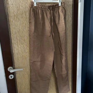 Bruna linnebyxor med snörning - Säljer ett par bekväma och stilrena bruna linnebyxor. De har en avslappnad passform med snörning i midjan för extra komfort. Perfekta för en avslappnad dag eller en somrig utflykt.