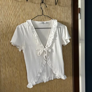 Vit kortärmad topp från NA-KD - Säljer en söt vit topp från NA-KD i storlek S. Den har korta ärmar och en v-ringning med volangdetaljer. Toppen är knytbar framtill och har en croppad passform. Perfekt för sommaren! 💕Har en liten fläck på ena snöret, men syns inte när man har på sig den.