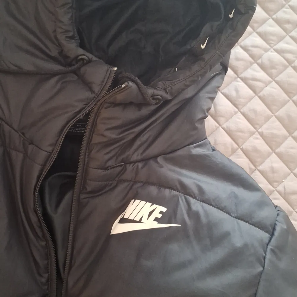 Säljer en svart pufferjacka från Nike i storlek XS. Jackan är perfekt för kalla dagar med sin vadderade design och har en dragkedja framtill. Den har en huva för extra skydd mot väder och vind. Nike-loggan syns tydligt på bröstet och längs nederkanten, vilket ger en sportig look.. Jackor.