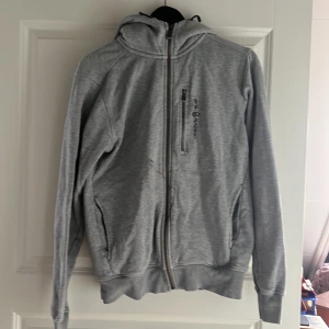 Grå hoodie från Sail Racing - Säljer en grå hoodie från Sail Racing i storlek S. Den är använd 3 gånger men fortfarande i väldigt bra skick. Gar däremot en defekt som spillts överdragkejdan vilken går att tvätta bort med starkare medel. Ordinare pris 1500kr nypris 800kr pga defekt