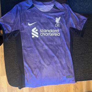 Lila Liverpool fotbollströja med M. Salah 11 - Säljer en snygg lila Liverpool fotbollströja från Nike. Den har ett coolt mönster och är prydd med 'Standard Chartered' loggan på framsidan. På baksidan står det 'M. Salah' och nummer 11. Tröjan är i nyskick och perfekt för alla Liverpool-fans!