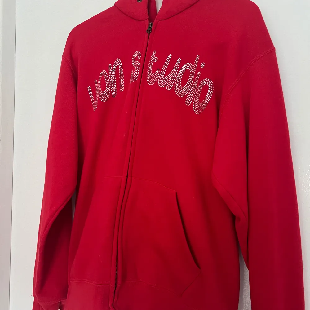 Säljer en supercool röd hoodie med dragkedja och strassdetaljer från Von Studio. Den har en glittrig text på framsidan och ett snyggt mönster på baksidan. Perfekt för att sticka ut i mängden! 🛍️. Tröjor & Koftor.