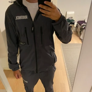 Grå vindjacka och vindbyxor från Hoodrich - Säljer en grå windbreaker samt tillhörande byxor från Hoodrich i storlek M. Perfekt för vår och höst. Jackan och byxorna är i nyskick och har en normal passform. De är tillverkad i polyester och är väldigt bekväma att ha på sig.