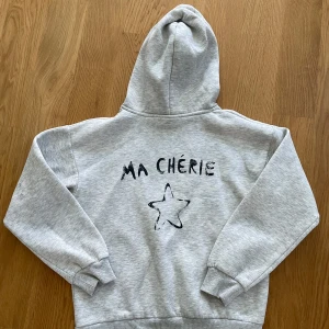 Grå hoodie från Gina Tricot - Säljer en supermysig grå hoodie från Gina Tricot med texten 'MA CHÉRIE' och en stjärna tryckt både fram och bak. Perfekt för en chill dag eller när du vill känna dig extra bekväm. Den har en klassisk passform med långa ärmar och en skön huva.