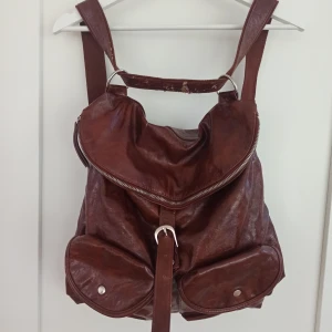 Brun ryggsäck från Complement - Säljer en snygg brun ryggsäck/axelväska från Complement. Den är gjord av syntet läder och har två justerbara axelremmar. Väskan har en dragkedja upptill och två fickor med knappar på framsidan🌟 Perfekt för vardagsbruk eller utflykter! enda defekten är att lädret har börjat flagna av på en del av axelremmen. 