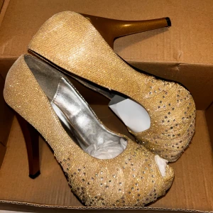 Glittriga guldiga pumps från Amar Shoes - Säljer ett par glittriga guldiga pumps från Amar Shoes. De har en elegant peep-toe design och hög klack, perfekt för festliga tillfällen. Skorna är dekorerade med små glittrande stenar som ger en extra touch av glamour.