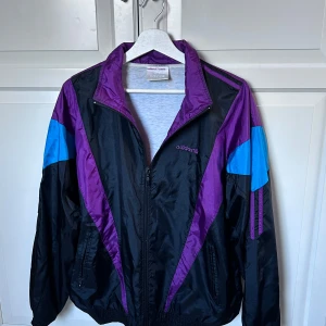 Svart och lila vindjacka från Adidas - Säljer en retro svart vindjacka från Adidas med lila och blå detaljer. Jackan har en dragkedja framtill och en klassisk krage. Strl S/M.  Perfekt för en sportig look eller en avslappnad dag ute. Hör av er vid intresse/ frågor