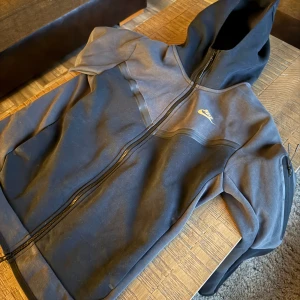 Svart och grå hoodie från Nike - Säljer en svart och grå hoodie från Nike med dragkedja och huva. Den har en guldig Nike-logga på bröstet och är tillverkad i ett mjukt material. Perfekt för en casual look eller träning.