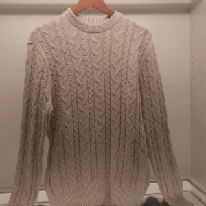 Beige stickad tröja från 157 - Säljer en snygg beige stickad tröja från 157. Tröjan har ett klassiskt flätmönster och ribbade muddar vid hals, ärmslut och nederkant. Perfekt för kyliga dagar!