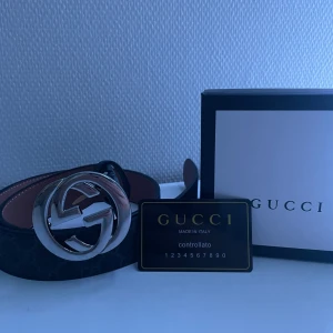 Svart bälte från Gucci - Snyggt svart bälte från Gucci med ett stort, glänsande silverfärgat spänne i form av Gucci-logotypen. Bältet har ett diskret mönster och är tillverkat i skinn. Perfekt för att ge en lyxig touch till din outfit. Skriv om du har några funderingar. Säljer pga för liten