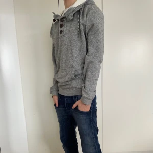 Grå hoodie med knappar - Säljer en stilren grå hoodie med knappar och snörning i halsen. Tröjan har en mysig huva och en praktisk magficka. Perfekt för kyliga dagar! Skick 9/10. Modellen är 177.