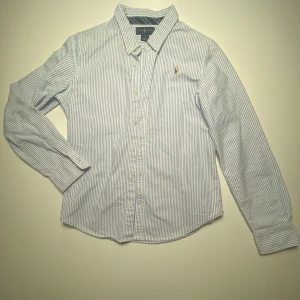 Randig skjorta från Ralph Lauren - Snygg vit och blå randig skjorta från Ralph Lauren med broderad logga på bröstet. Skjortan har långa ärmar och knappar framtill. Perfekt för en stilren look. Passar dig mellan 150-160. Priset kan diskuteras!