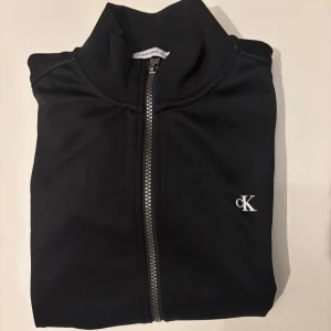 Calvin Klein svart tröja - Snygg svart tröja från Calvin Klein Jeans med dragkedja och hög krage. Den har en stilren design med CK-logga på bröstet och ribbade muddar. Perfekt för en stilren look. Mycket bra skick. Köps för 699kr med tillhörande byxor. Barnstorlek, 160.