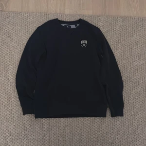 Mörkblå sweatshirt från GANT - Säljer en stilren mörkblå sweatshirt från GANT med ett broderat emblem på bröstet. Tröjan har långa ärmar och en klassisk rund halsringning. Perfekt för en avslappnad och snygg look.