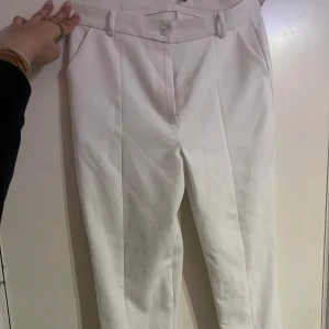 Vita kostymbyxor - Snygga vita kostymbyxor med hög midja och knappdetalj. Perfekta för en stilren look. Byxorna har en klassisk design med raka ben och bälteshällor.