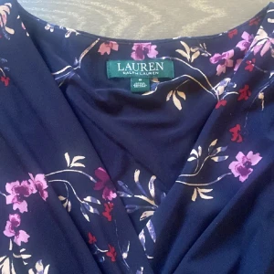 Blommig klänning från Ralph Lauren - Snygg blommig klänning från Ralph Lauren i mörkblått med rosa och lila blommor. Klänningen har en omlott-design och är ärmlös, vilket ger en elegant look. Perfekt för en somrig dag eller en speciell kväll.