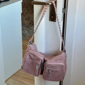 Rosa axelväska med nitar - Snygg rosa axelväska i mocka med flera dragkedjefickor och coola nitar på axelremmen. Perfekt för att ge din outfit en edgy touch. Justerbar rem för bekväm passform.