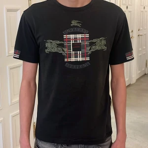 Svart t-shirt från Burberry - Snygg svart t-shirt från Burberry med klassiskt rutmönster och hästmotiv. T-shirten har korta ärmar med rutiga detaljer och ett tryck på bröstet som ger en stilren look. Det är en vintage tröja som jag tror man inte kan köpa längre fick den av min farfar