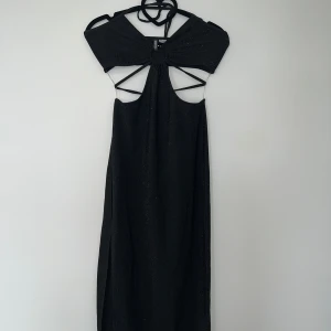 Svart glittrig klänning med öppen rygg - Snygg svart klänning med glittrigt tyg och öppen rygg. Den har en unik design med snörning och knytning i ryggen. Perfekt för en kväll ute eller en speciell tillställning.