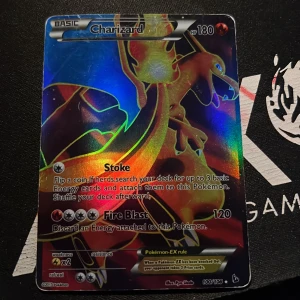 Charizard Pokémon-kort - Säljer ett Charizard Pokémon-kort med holografisk yta. Kortet har 180 HP och innehåller attacker som Stoke och Fire Blast. Det är en del av en samling och har nummer 100/103. Perfekt för samlare eller Pokémon-entusiaster!