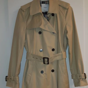Beige trenchcoat från Mango - Snygg beige trenchcoat från Mango aldrig använt, med klassisk dubbelknäppning och bälte i midjan. Jackan har långa ärmar och ett stilrent rutigt foder. Perfekt för höst och vår!