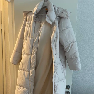 Beige dunjacka med huva - Säljer en lång beige dunjacka med huva. Jackan har en dragkedja framtill och långa ärmar. Perfekt för kalla dagar när du vill hålla dig varm och bekväm. Hör av dig vid intresse! 