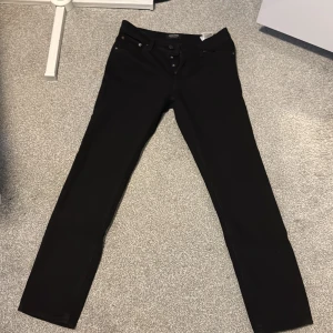 Svarta jeans från Jack & Jones - Snygga svarta jeans från Jack & Jones med knappar i midjan. Perfekta för en stilren look. De har en klassisk design med fem fickor och en smal passform.