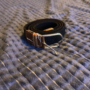 Svart bälte med brunt läderdetalj - Snyggt svart bälte med en stilren brunt läderdetalj vid spännet. Bältet har ett klassiskt silverfärgat spänne som ger en tidlös look. Perfekt för att komplettera din outfit.
