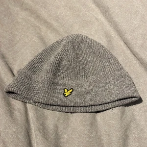 Grå stickad mössa från Lyle & Scott - Snygg grå stickad mössa från Lyle & Scott med deras ikoniska gula logga framtill. Perfekt för kyliga dagar med sin ribbade design som ger en skön passform. En stilren accessoar för både höst och vinter.