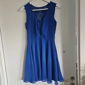 Blå ärmlös klänning med spetsdetaljer - Snygg blå klänning med v-ringning och ärmlös design. Baksidan har vackra spetsdetaljer med broderade blommor. Perfekt för en somrig look. Ska vara med ett skärp i midjan som gått sönder, så finns hällor på sidorna för plats till ett smalt skärp. 